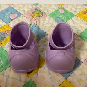 Pode incluir: Um par de sapatos de boneca de plástico lavanda com um laço na frente. Os sapatos estão em um tecido acolchoado com um padrão colorido.