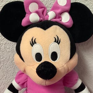 May include: A Minnie Mouse soft toy, with a pink dress and white polka dots, and a matching bow. The toy has black ears, a black nose, and white gloves. The toy is sitting on a pink surface.