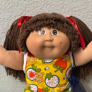May include: A vintage doll with brown yarn hair styled in pigtails, adorned with red ribbons. The doll wears a yellow dress with a strawberry and teddy bear pattern, green shorts, white socks, and white shoes with lace trim. A blue bag hangs from the doll's arm.