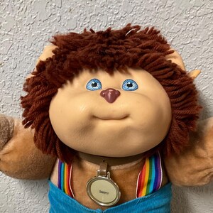 Könnte beinhalten: Ein Stofftier mit rundem Gesicht, blauen Augen und braunen Haaren. Es trägt blaue Cordshorts mit Regenbogen-Hosenträgern und einem Anhänger mit der Aufschrift "Dappy". Das Spielzeug sitzt auf einem gemusterten Quilt.