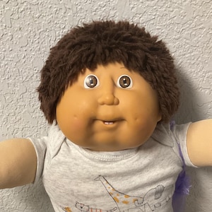 May include: A vintage doll with brown hair and large, expressive eyes. The doll wears a gray onesie with animal graphics, gray pants, and white sneakers with blue stripes. The doll is sitting on a colorful quilt.