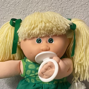 May include: A vintage Cabbage Patch Kids doll with blonde yarn hair styled in pigtails, green eyes, and a pacifier. The doll wears a green dress and shoes, with white lace trim. The doll is sitting on a colorful quilt.