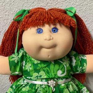 May include: A vintage Cabbage Patch Kid doll with red yarn hair styled in pigtails with green bows. The doll wears a green floral dress with a white ribbon belt and white shoes. The doll has blue eyes and a smiling expression.