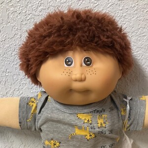 May include: A Cabbage Patch Kid doll with brown hair, freckles, and large eyes. The doll wears a gray shirt with yellow tiger prints and yellow pants. The doll is sitting on a colorful quilt.