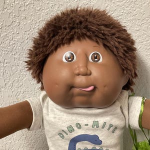 Pode incluir: Boneca Cabbage Patch Kid com pele castanha, cabelo castanho e expressão brincalhona. A boneca usa uma camiseta cinza com um gráfico de dinossauro azul e as palavras "Dino-Mite Little Dude", calças azuis e sapatos brancos.