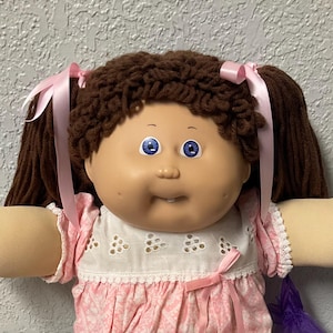 May include: A vintage Cabbage Patch Kid doll with brown yarn hair styled in pigtails, blue eyes, and a rosy complexion. The doll wears a pink dress with white lace trim, white socks, and white shoes. A purple bag is visible.