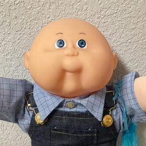 May include: A vintage Cabbage Patch Kid doll with a bald head and blue eyes. The doll is wearing a blue plaid shirt, denim overalls, and white sneakers. The overalls have gold-colored clasps.