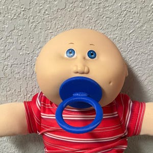 May include: A Cabbage Patch Kids doll with a bald head, blue eyes, and a blue pacifier. The doll is wearing a red and white striped shirt, blue denim pants, and white socks. The doll's arms are outstretched.