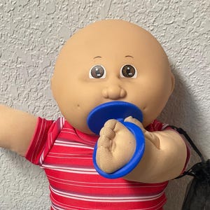 May include: A Cabbage Patch Kids doll wearing a red and white striped shirt, blue denim jeans, and a blue pacifier. The doll has a bald head and is holding a black mesh bag.