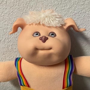 May include: A plush doll with a round peach-colored face, brown eyes, and white yarn hair. The doll features floppy ears, a small brown nose, and wears yellow corduroy shorts with rainbow-striped suspenders and a small white graphic on the hip.