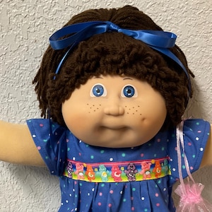 May include: A doll with dark brown yarn hair tied with a blue ribbon, blue eyes, and freckles. It wears a blue dress with colorful polka dots and a ribbon featuring cartoon bears, white socks with ruffled lace trim, and a small pink organza bag.
