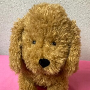 May include: A plush, golden-brown dog toy with a fluffy texture. The toy has a black nose, small black eyes, and floppy ears. It is sitting on a pink surface.