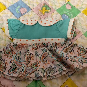 May include: A doll dress featuring a teal bodice with a white collar and cuffs accented with orange polka dots. The skirt is a paisley print in pink, orange, and blue. The dress is displayed on a colorful quilt.
