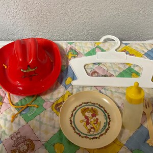 May include: A collection of vintage Cabbage Patch Kids toys, including a red cowboy hat with a gold star, a plate, a bottle, a fork, a spoon, and a white hanger. The items are arranged on a colorful quilted mat.