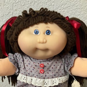 May include: A vintage doll with brown yarn hair styled in pigtails, adorned with red ribbons. The doll wears a gray floral dress with a white lace trim and pink socks. A white cloth bag hangs on the side.