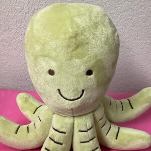 May include: A green plush octopus with a smiling face and brown embroidered details. The octopus has eight tentacles.