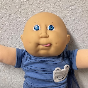 Op de afbeelding: Een Cabbage Patch Kids pop met een kale kop, blauwe ogen en een speelse uitdrukking. Het draagt een blauw gestreept shirt met een kikkerzak, een zwarte broek en witte sneakers met blauwe accenten. Een blauwe, doorschijnende cape is bevestigd.
