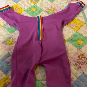 May include: A purple jumpsuit with rainbow trim on the sleeves and zipper. The jumpsuit is laid on a colorful quilted blanket with cartoon characters and balloons.