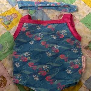 May include: A blue swimsuit and matching headband set. The swimsuit has a wave pattern with pink and white accents, and pink trim. The headband mirrors the swimsuit's design. The set is designed for a doll.
