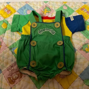 May include: Green and yellow Cabbage Patch Kids dungarees with blue trim and buttons. The dungarees have a white elastic waistband and are made of a soft, cotton fabric.
