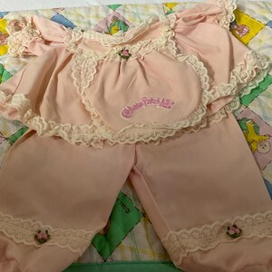 May include: A pink baby doll outfit with lace trim and a flower embroidered on the front. The outfit says "Cabbage Patch Kids" on the front.
