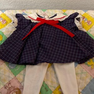 May include: A vintage navy blue plaid dress with a white collar and red bow. The dress has white lace trim and is paired with white tights. The outfit is displayed on a colorful patchwork quilt.