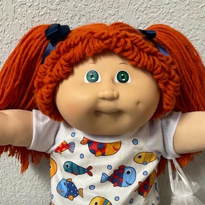 May include: A Cabbage Patch Kid doll with orange yarn pigtails tied with blue ribbons. The doll wears a white shirt with a colorful fish pattern, white shoes, and a white lace trim. The doll has green eyes and rosy cheeks.
