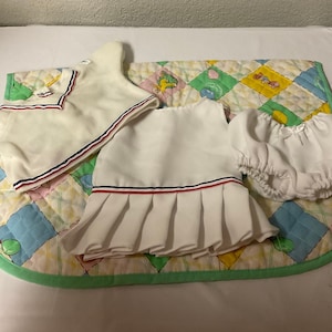 May include: A white baby dress with red and blue trim, a white sweater with red and blue trim, and a white diaper with a bow. The items are laid out on a green and yellow patterned fabric.