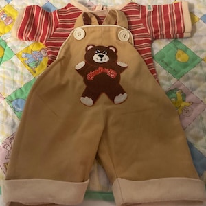 May include: A vintage baby outfit featuring a tan overall with a teddy bear applique and a red and white striped long-sleeved shirt. The overall has white trim and large buttons. The outfit is displayed on a colorful quilt.