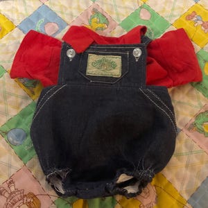 May include: A vintage doll outfit featuring a red top with puffed sleeves and a denim blue overall. The overall has white stitching and two button closures on the straps. A rectangular patch with text is sewn on the front. The outfit is on a colorful quilt.