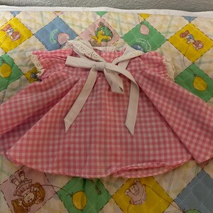 May include: A vintage pink and white gingham dress with a white bow and collar. The dress has short sleeves and a flared skirt. The dress is likely for a baby or toddler. The dress is on a colorful quilt.