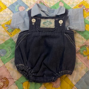 May include: A dark blue denim overall romper with white stitching and two white buttons at the straps, layered over a light blue and white gingham short-sleeved collared shirt. A rectangular Cabbage Patch Kids label is sewn onto the chest pocket.
