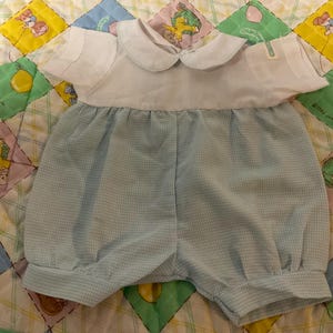 May include: A baby's romper set with a white top and light blue checkered shorts. The top has a rounded collar and short sleeves. The shorts are gathered at the waist and legs. The romper is placed on a colorful quilt.
