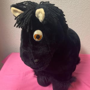 May include: A plush black horse puppet with a detailed mane and large, expressive eyes. The puppet has a soft, furry texture and is set against a pink background. The horse's ears are a light tan color.