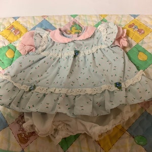 Könnte beinhalten: Ein hellblaues und rosa Baby-Puppenkleid mit weißem Spitzenbesatz und einem Blumenmuster. Das Kleid hat einen rosa karierten Kragen und kurze Ärmel.