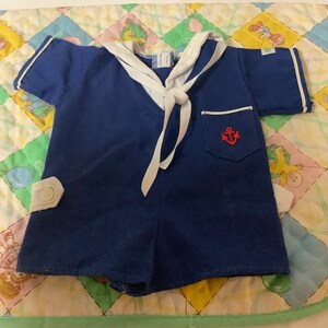 May include: A blue sailor-style baby onesie with a white collar and a red anchor on the pocket. The onesie has a white tie at the neck.