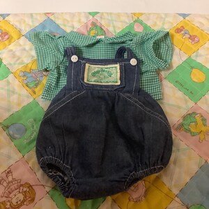 May include: A Cabbage Patch Kids outfit for a doll. The outfit includes a green and white gingham shirt and blue denim dungarees with a Cabbage Patch Kids label.
