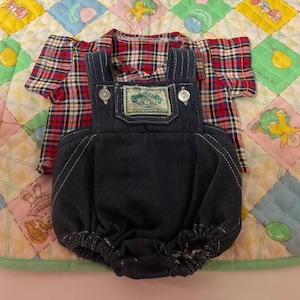 Peut inclure: Une chemise à carreaux rouge et bleu et une salopette en jean avec une étiquette "Cabbage Patch Kids". La tenue est posée sur une couverture matelassée colorée.