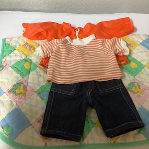 Può includere: Un set di vestiti per una bambola, che comprende una maglietta a righe, pantaloncini di jeans blu e una giacca arancione brillante.