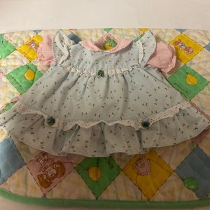 Puede incluir: Un vestido de muñeca azul claro y rosa con ribete de encaje blanco y estampado floral. El vestido tiene un cuello y mangas de vichy rosa.