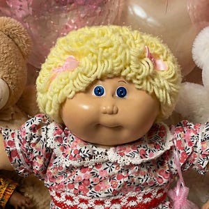 May include: A central Cabbage Patch Kids doll with yellow yarn hair and a floral dress, surrounded by four smaller dolls in diverse outfits, two plush teddy bears, heart-shaped balloons, chocolate chip cookies, brownies, and pink candies.
