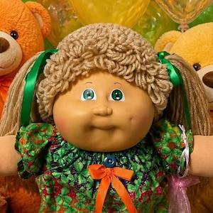 May include: A Cabbage Patch Kids doll with yarn hair and a green patterned dress sits among several smaller baby dolls, two teddy bears, plates of chocolate chip cookies and brownies, and bowls of colorful jelly beans and gummy candies.