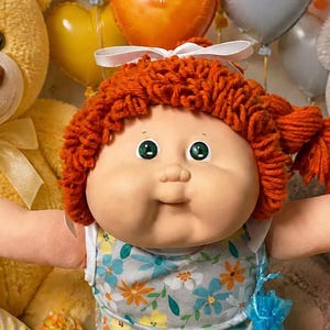 May include: A doll with orange yarn hair and green eyes wearing a floral dress, white socks with lace trim, and white shoes, surrounded by plush teddy bears, baby dolls, heart-shaped balloons, cupcakes, cookies, and wrapped candies.