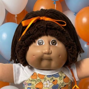 Puede incluir: Una muñeca grande con cabello de lana marrón y un lazo naranja, que viste una blusa blanca y amarilla con estampado de fresas, calcetines blancos con encaje y zapatos blancos. Cuatro muñecas más pequeñas se sientan sobre una colcha.