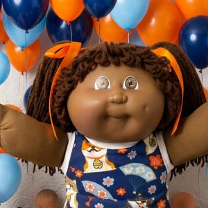 May include: A dark-skinned doll with brown yarn hair in pigtails tied with orange ribbons, wearing a blue sleeveless dress featuring a cat and fan pattern, white ruffled socks, and black and white shoes, surrounded by various smaller dolls and teddy bears.