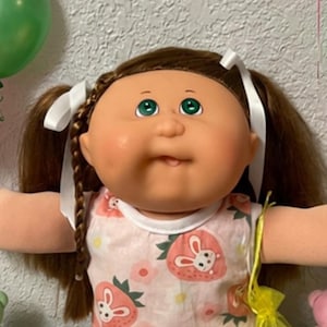 May include: A Cabbage Patch Kids doll with brown yarn hair in pigtails tied with white ribbons, green eyes, and a light skin tone. The doll wears a sleeveless white dress with a pink strawberry and rabbit print, white socks with lace trim, and white shoes.