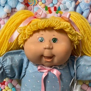 May include: A large doll with yellow yarn hair in pigtails, wearing a blue dress with white patterns and a pink ribbon, surrounded by smaller dolls, light blue teddy bears, colorful balloons, lollipops, and various small candies.