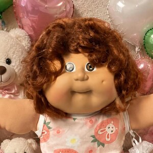 May include: A doll with curly auburn hair and a round face wearing a sleeveless white top featuring a pink strawberry and bunny print, white socks with ruffled lace trim, surrounded by various plush teddy bears, heart balloons, and cookies.