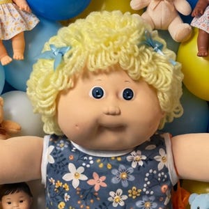 May include: A large doll with curly yellow yarn hair and blue eyes wearing a blue floral top and ruffled socks, surrounded by smaller dolls, miniature teddy bears, yellow and blue balloons, and wrapped candies scattered across the background.