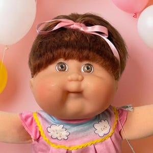 May include: A large doll with brown hair and a pink headband, wearing a pink patterned top, sits surrounded by seven smaller dolls of various ethnicities in colorful outfits. The scene is decorated with pink, yellow, and white balloons.